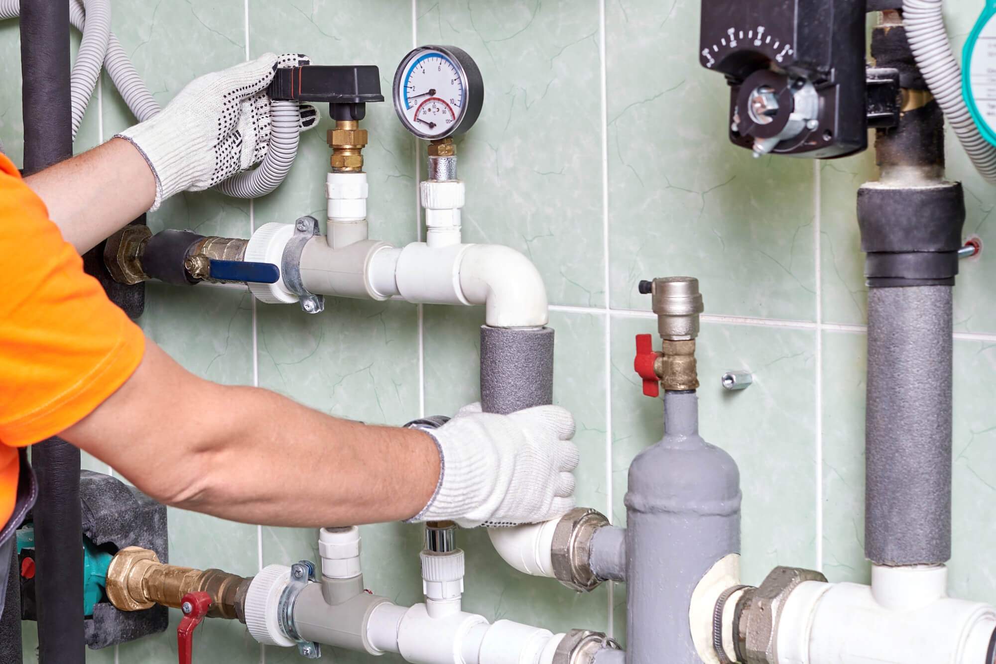 A plumber in an orange shirt and white gloves working on a commercial plumbing system with various pipes, valves, and gauges.